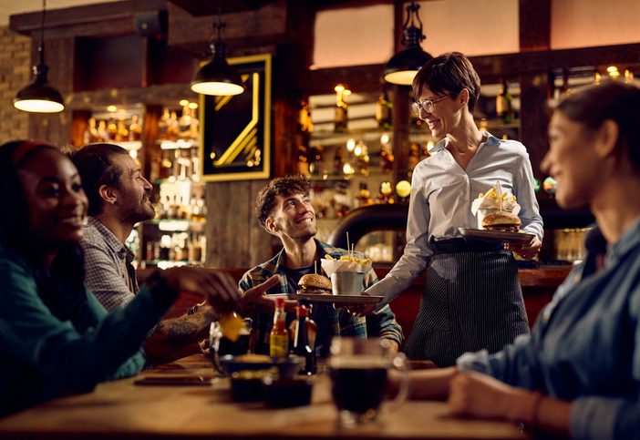 Happy waitress serving hamburger with French fries to group of guests in a bar.