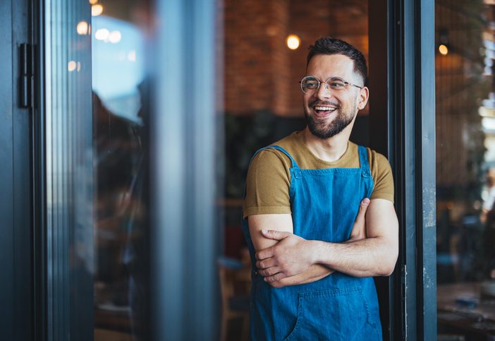Smiling male bartender, clad in apron, stands arms crossed in a cozy coffee house, ready to serve a diverse clientele with a variety of coffee beverages.